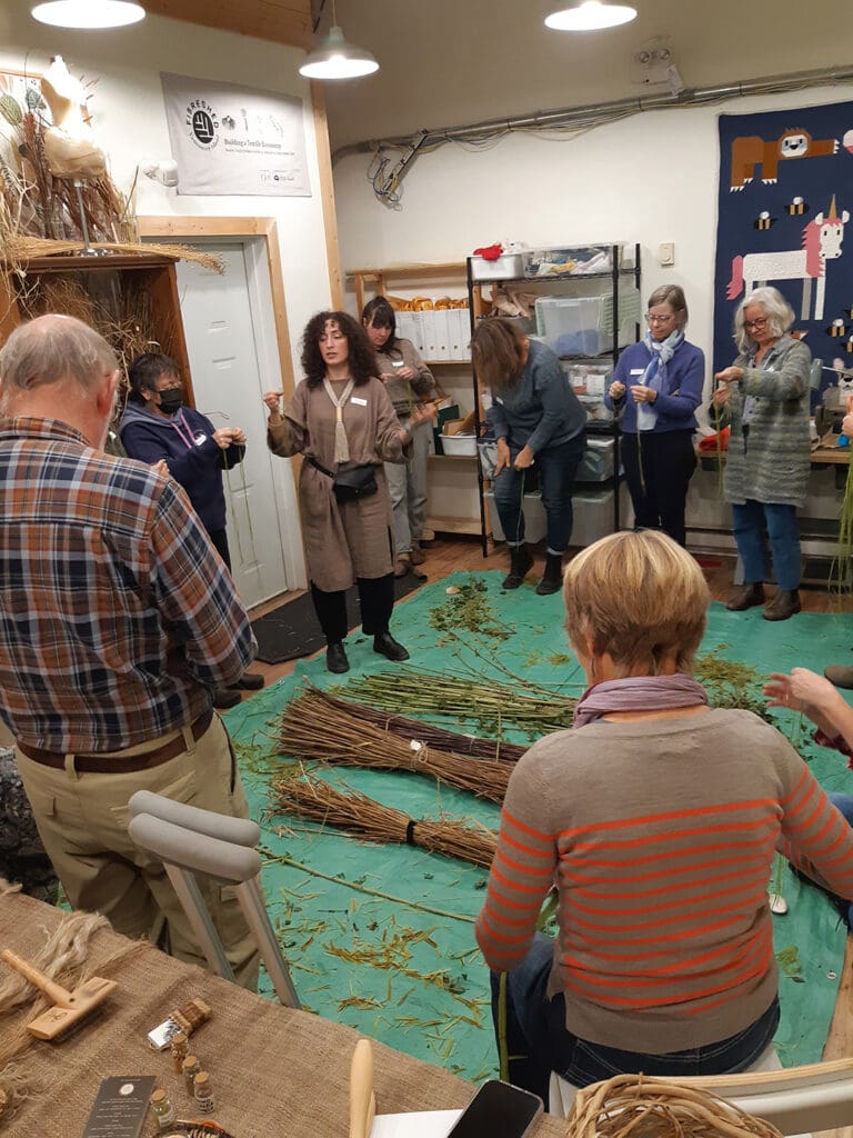 people standing around a green tarp on the floor of a studio room, watching Juliana describe how to process nettle fibres