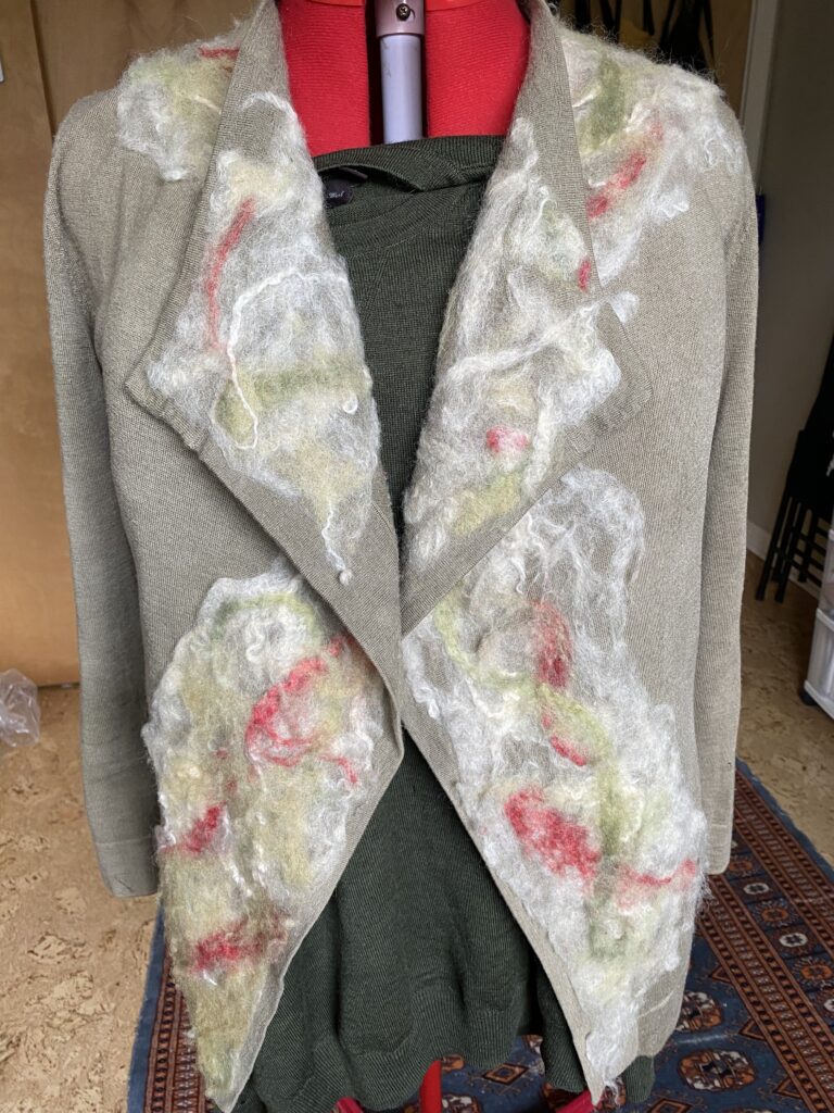 front view of a olive green/grey cashmere cardigan with wool fibres in white, green, and red needle-felted to the lapels, draped over a red dress form. 