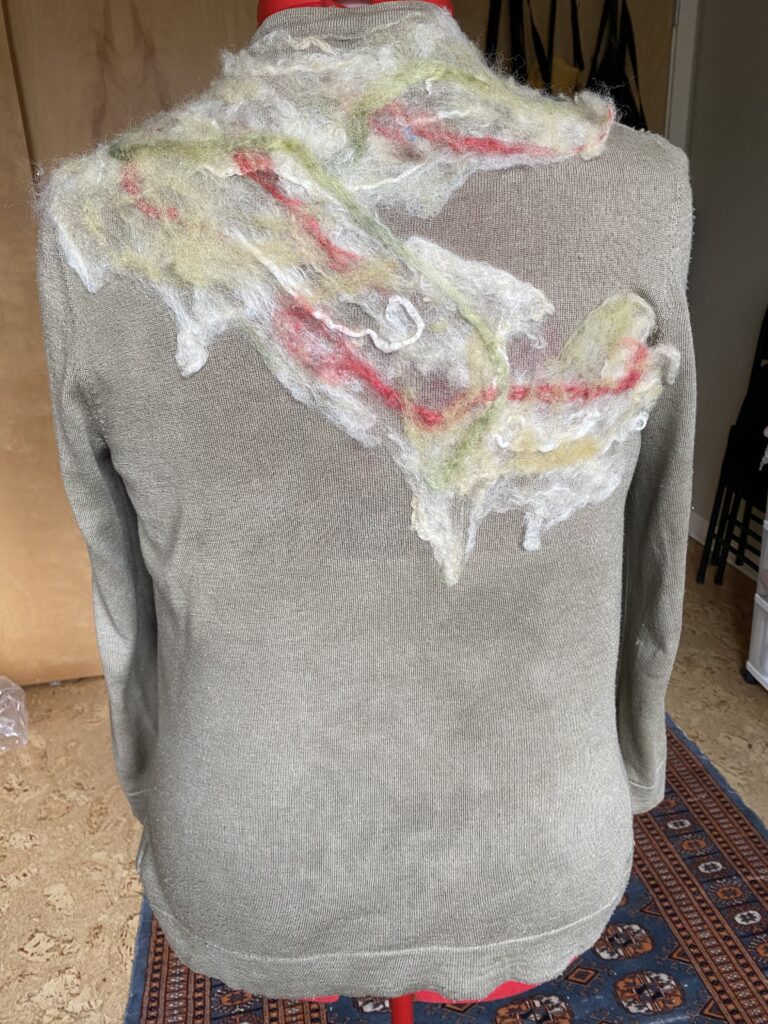 back view of a olive green/grey cashmere cardigan with wool fibres in white, green, and red needle-felted to the top shoulder area, draped over a red dress form. 