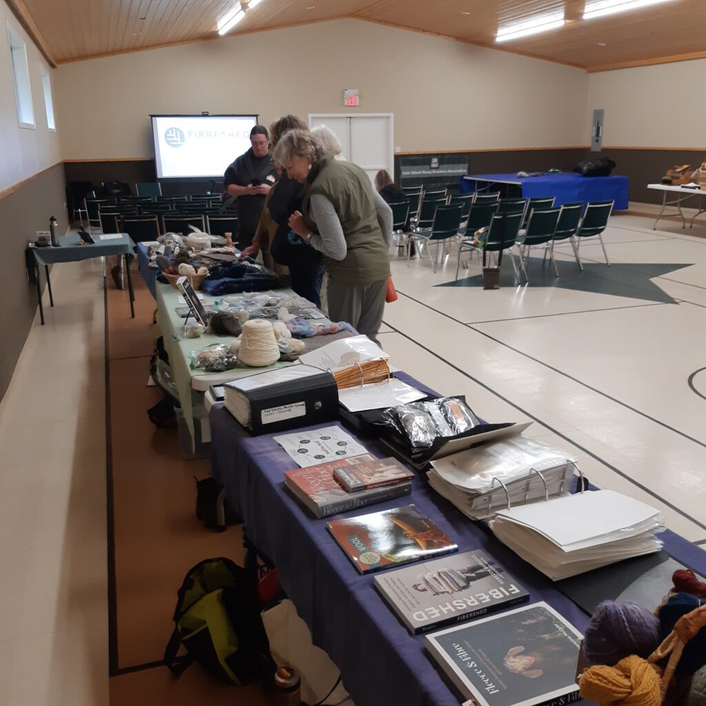 people looking at books and yarn samples on a table in a gym. 