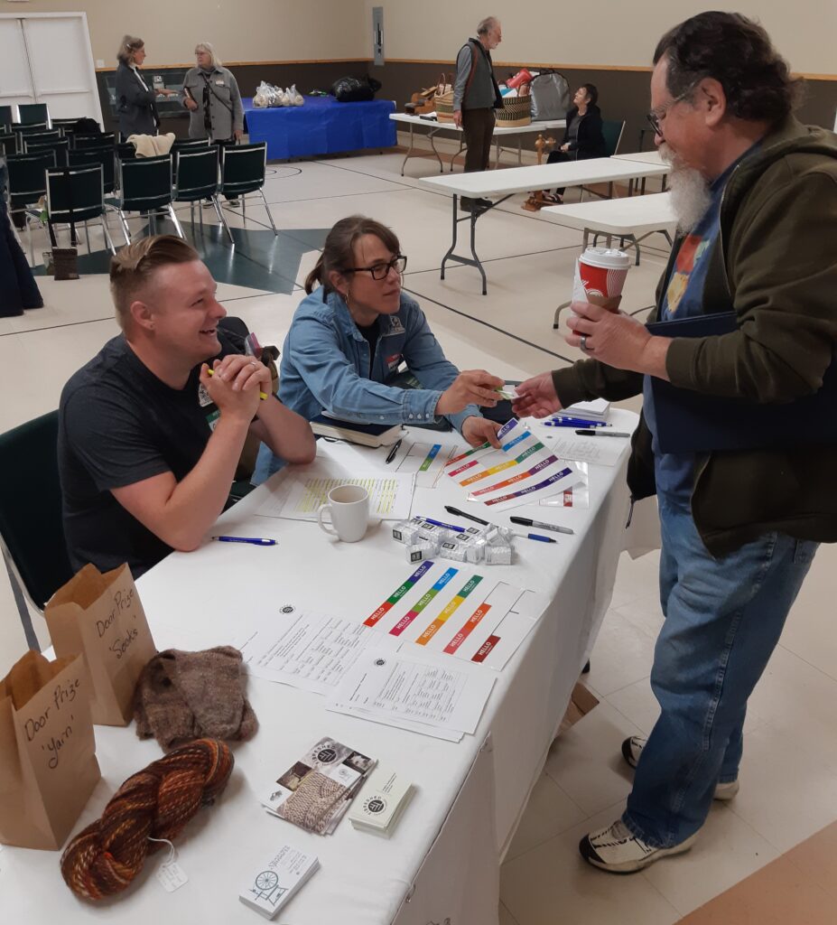 two people at a registration table handing a nametag to a third person, holding a coffee. Colourful yarn skein and handknit socks are offered as door prizes. 