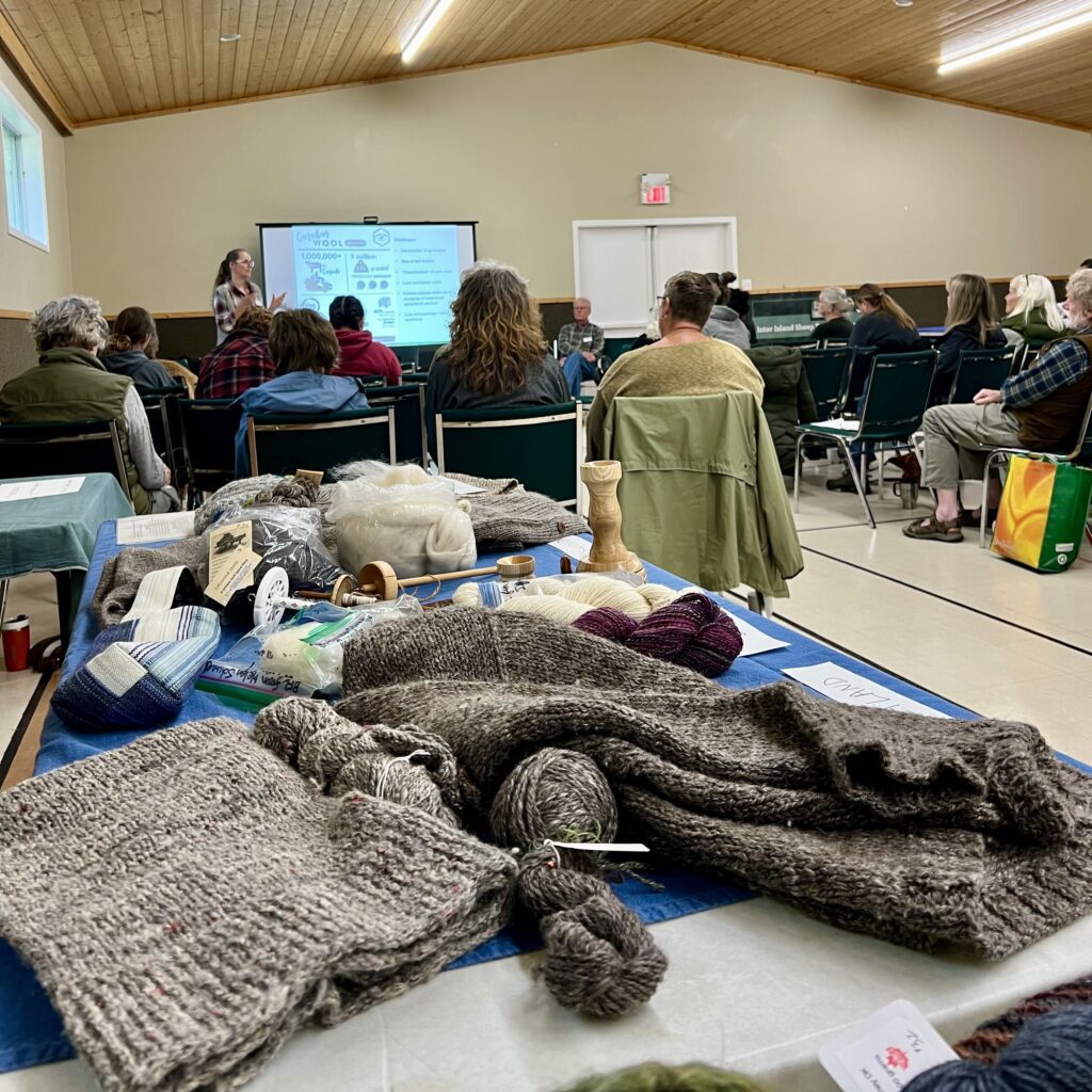 In the foreground, handknit grey/brown knitted fabric, including vest and sweater, with the yarns used to make them, on a table. In the background, people in chairs listening to a woman presenting a slide presentation on a screen in a gymnasium. 