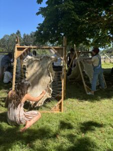 a woman with her back to the camera working on a hide stretched on a wooden frame. Green grass, blue sky.