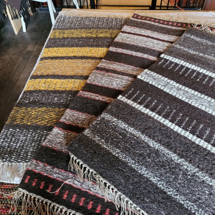 three stacked rugs on a wood table. Rugs are boundweave (weft faced) in natural sheep shades and yellow and orange naturally dyed yarn. 