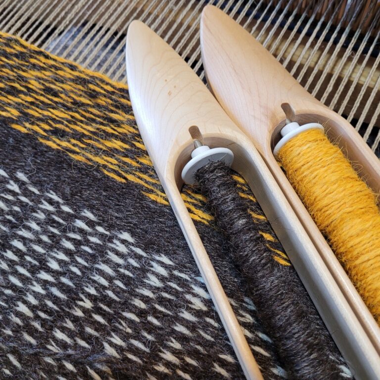 close up of weaving in progress showing linen warp and brown, white, and yellow wool yarns. Two wood shuttles sit on top of the woven fabric, with brown and yellow wool on the bobbins in the shuttles.