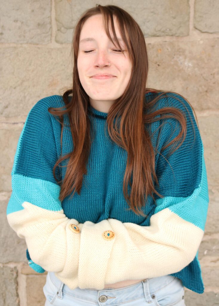 vibrant sweater in cream, light blue, and dark teal, worn by a smiling white woman with long dark hair. Her arms are crossed and her eyes are closed - she's giving herself a hug.