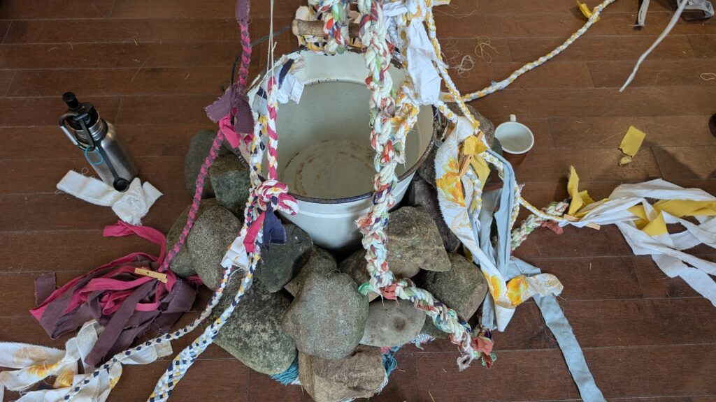 white metal bucket surrounded by stones on wood floor. Braided cordage hangs around the bucket. 