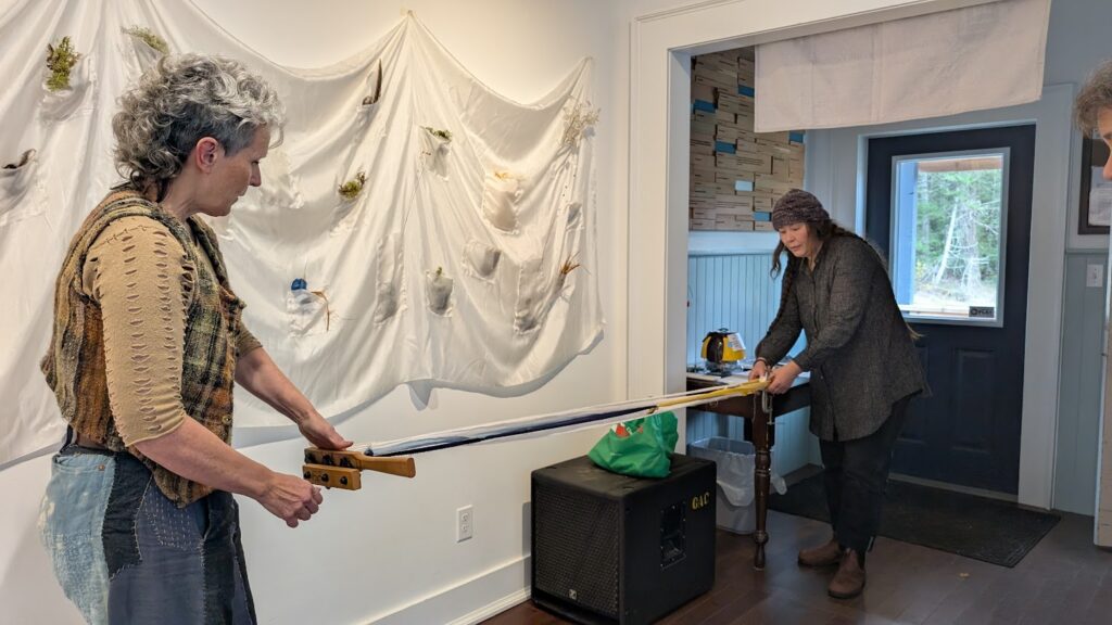 two women (Sharon and Yarrow) work together to tension fabric strips. In the background is  a white silk cloth panel with pockets containing fragments of Gabriola landscape hanging on the wall. 